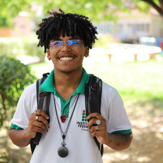 Estudante sorridente com a camisa do uniforme do Instituto Federal do Ceará e mochila nas costas, em área arborizada de um campus